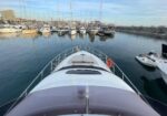 View from the bow of a white yacht looking into a marina filled with moored boats and sailboats, calm water reflecting the vessels and a breakwater on the right.