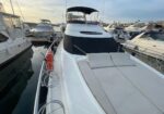 White luxury motor yacht docked in a marina with cushioned sun loungers on the bow and a dark window cover at the front.