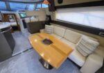 Salon of a luxury yacht: curved cream sofa around a polished wooden table with built‑in hatch, beside large window blinds and a helm area nearby.