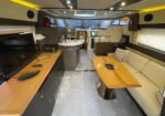 Luxurious yacht interior: beige sofa seating, wooden dining table, and a flat-screen TV beside a galley area near the helm point.
