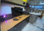 Luxurious camper-van interior with a flat-screen TV on a wooden counter, remote, and small work area including a printer and wireless device, under purple ambient lighting.