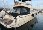 White motor yacht docked at a marina with a black canvas cabin, wooden swim platform, and fenders along the hull.
