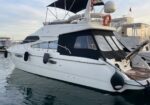 White motor yacht docked at a marina with black cockpit canvas and covered windows; calm water around it and other boats in the background.