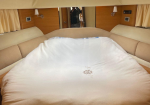 A large bed in a beige yacht cabin with curtains and wooden walls; white blanket with a ship wheel embroidery covers the bed.