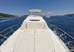Bow of a luxury white yacht with padded sun loungers, railing on both sides, sailing on blue sea with distant boats and coastline on the left.