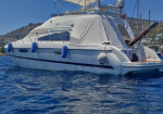 White motor yacht floating on blue water with fenders along the hull and a shaded aft deck, hillside town in the background.