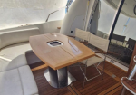 Oval wooden table with a built-in cup holder on a yacht deck, surrounded by cream cushioned seating and folding chairs