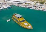 A large yellow passenger yacht docked in a marina with numerous smaller boats along the pier and teal water surrounding it.