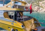 Yellow and gray motor yacht with an open rear deck dining area, turquoise water and a rocky coastline in the background, a man standing inside.