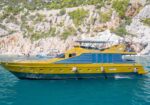 Yellow and blue luxury motor yacht anchored near rocky cliffs with turquoise water and clear sky behind it.