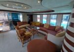 Luxurious yacht salon with brown and beige sectional sofas, wicker chairs, and a glass-topped coffee table overlooking an outdoor deck and pool area.