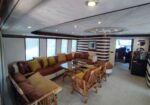 Luxurious yacht salon with a curved brown sofa, colorful cushions, wicker chairs, and a glass coffee table near striped walls.