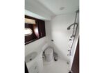 Compact white bathroom with toilet, sink, and vertical shower rail; porthole window and dark wood cabinet above sink.