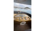 View from the yacht cockpit: gold control panel with gauges, steering wheel, and nav gear overlooking a marina with boats and a small island in the distance.