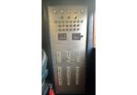 Industrial electrical control panel with many circuit breakers and green indicator lights, two digital meters at top, in a utility room.