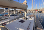 Sailing boat docked at a marina with sails lowered, rigging visible and calm blue water in the background.