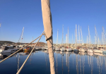 View from a sailboat deck toward a marina with numerous moored sailboats and their tall masts reflected in the calm water, foreground wrapped mast fittings visible.