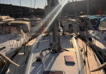 Deck view of a sailboat in a marina at sunset, with rigging, winches, and hatches visible on a wooden deck.