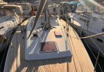 White sailing yacht docked in a marina, showing a wooden deck, hatch covers, and surrounding boats