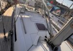 View along the deck of a white sailing yacht at a sunny marina, with rigging and other boats nearby.