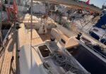 Sailing sailboat at a marina, view of the aft cockpit with coiled lines, winches, and a sunlit wooden deck under a clear sky.
