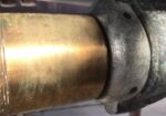 Close-up of a brass pipe secured by a gray metal clamp with holes, outdoors.