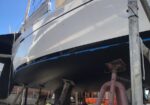 Sailboat on dry dock supported by metal stands, hull facing the camera with a blue sky behind.