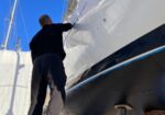 Worker in black clothing stands on a metal platform, inspecting the white sailboat hull supported on stands in a boatyard under a clear blue sky.