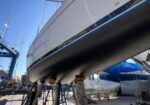 Large white sailboat on dry dock supports in a marina workshop, masts and scaffolding visible around it.