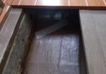 Open floor hatch on a wooden deck revealing a dark cavity with rusty metal and debris beneath the panel.