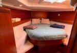 Cozy boat cabin with a round bed in the center, blue comforter, wrapped towels, and white cushioned seating along wooden walls under a skylight.