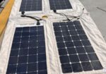 Four solar panels laid out on a white fabric mat at a dock, with cables and rigging nearby, likely for portable power.