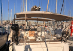 White sailboat docked in a sunny marina, beige cockpit canopy, steering wheel, and stern ladder visible among masts and neighboring boats (Turkish flag in background).