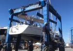 Blue high-capacity boat hoist supports a white sailing yacht in a shipyard; a worker stands on a platform beside the hull under a clear blue sky.
