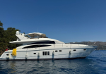 White luxury motor yacht anchored in calm blue water, with yellow mooring lines and distant coastline under a clear sky.