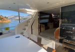 Open sliding door to a luxury yacht's interior lounge; deck area with a white table and seating, stairs leading to the upper deck, and calm blue water with rocky coast in the background.