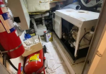 Cramped utility room with an open furnace/boiler, exposed panels and hoses, a red fire extinguisher on the left, and maintenance supplies on the floor.