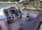 Boat cockpit interior with steering wheel, gauges, and Raymarine control panel by the water.