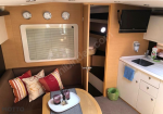 Compact yacht cabin: L-shaped sofa with striped cushions, coffee table, window with blinds, and a doorway to bunk berths; kitchenette with sink, soap, and a mounted TV nearby.