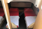 Two single beds with red duvets and patterned white sheets in a small cabin, separated by a narrow aisle.