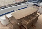 Wooden dining table with woven rattan chairs on a boat deck beside white cushioned seating.