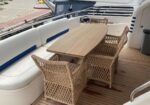Teak rectangular table with woven wicker chairs on a yacht deck, white cushioned seating along the side.