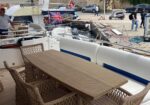 Yacht deck with a wooden dining table and wicker chairs, white cushioned seating, at a marina; people walk along the pier and a Union Jack flag waves on a nearby boat.