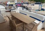Outdoor yacht deck with a wooden table and wicker chairs, white cushioned seating with blue accents, at a marina.