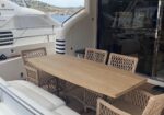 Outdoor dining area on a yacht deck: a rectangular wooden table surrounded by woven wicker chairs with the water and another boat in view behind.