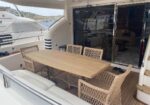 Outdoor yacht deck dining area with a wooden table and woven wicker chairs, next to glass sliding doors into the cabin.