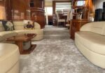 Luxurious yacht interior with curved beige sofas, wood paneling, and a carpeted salon area. A small glass-topped table sits between seating.