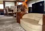 Cream curved sofa in a yacht's wood-paneled living area with a TV and dining nook in the background, plus a spiral staircase above.