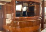 Glossy curved wooden bar inside a luxury yacht, with built-in shelves and a pass-through window to the helm area.