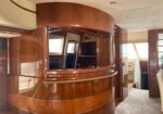 Luxurious yacht interior with glossy cherry wood paneling, a curved bar/desk, and navigation area visible through an opening in the wall.